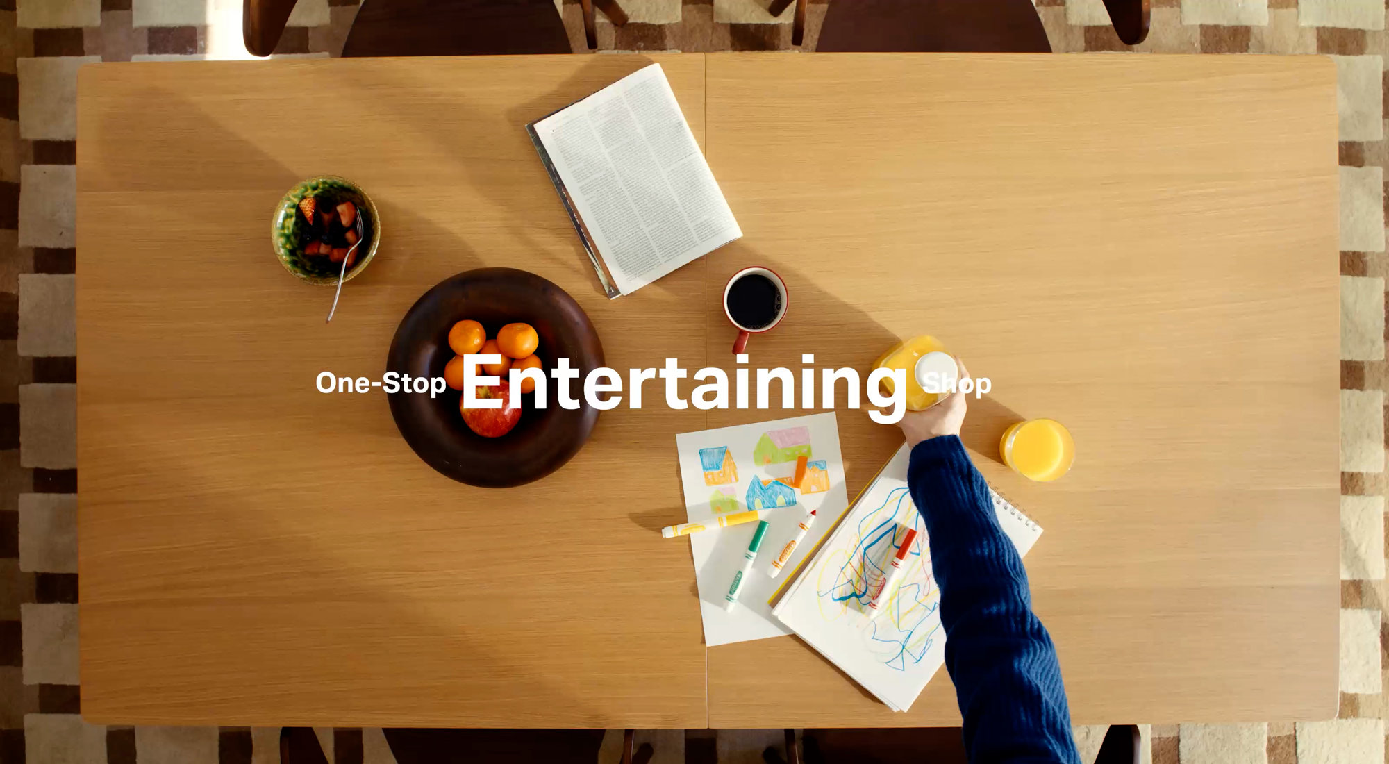 Overhead view of a light wood dining table with a bowl of fruit, coffee, a book, and art supplies, text reads One-Stop Entertaining Shop.