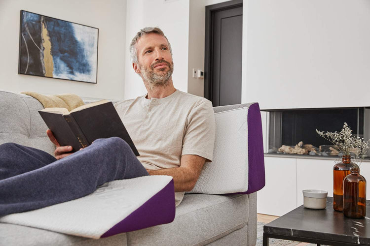 a man on a sofa with supportive pillows reading a book