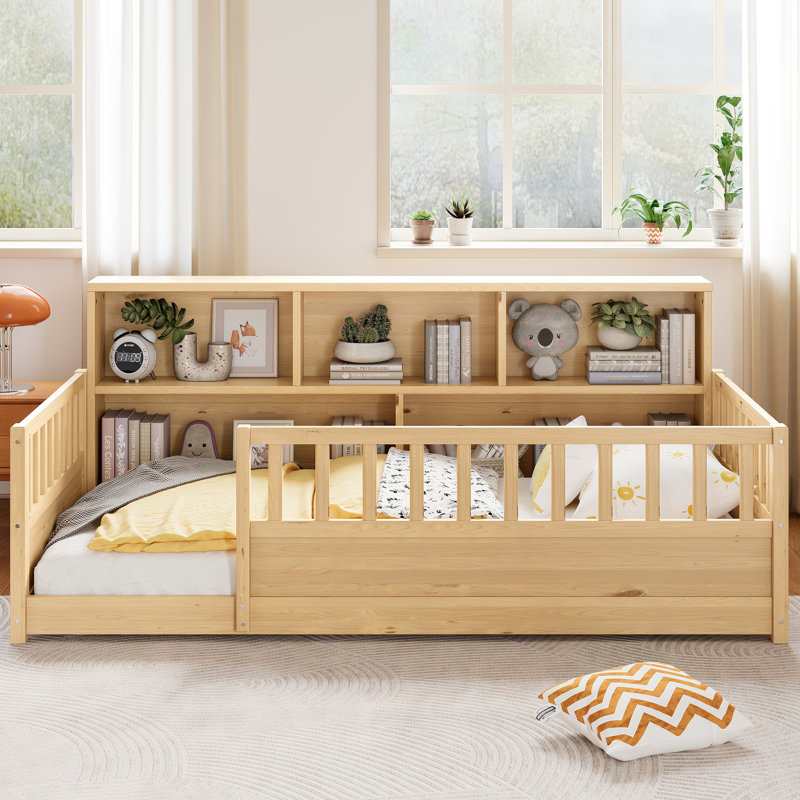 Un lit de plancher en bois simple avec une barrière de sécurité de style clôture, avec une fonction décorative de rangement latérale, et sa couleur est dans le ton de bois naturel.