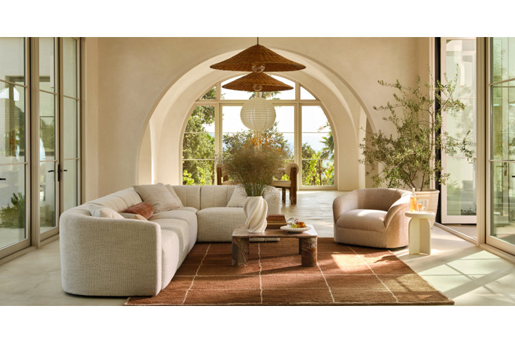 A serene Mediterranean-style living room framed by large arched doorways and floor-to-ceiling glass doors overlooking a lush outdoor landscape. The space features a low-profile cream bouclé sectional sofa and a matching rounded armchair resting on a rust-brown grid area rug. In the center sits a low marble coffee table decorated with a twisted white vase and a bowl of fruit. Tiered woven pendant lamps and a white paper lantern light hang from the ceiling, while a potted olive tree stands in the right corner.