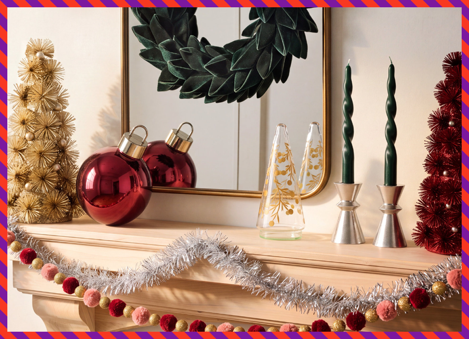 A festive fireplace mantel decorated with a gold starburst tree, a large red ornament, a glass tree, silver candle holders with dark green twisted candles, a red bottle brush tree, and a silver tinsel and pom-pom garland.