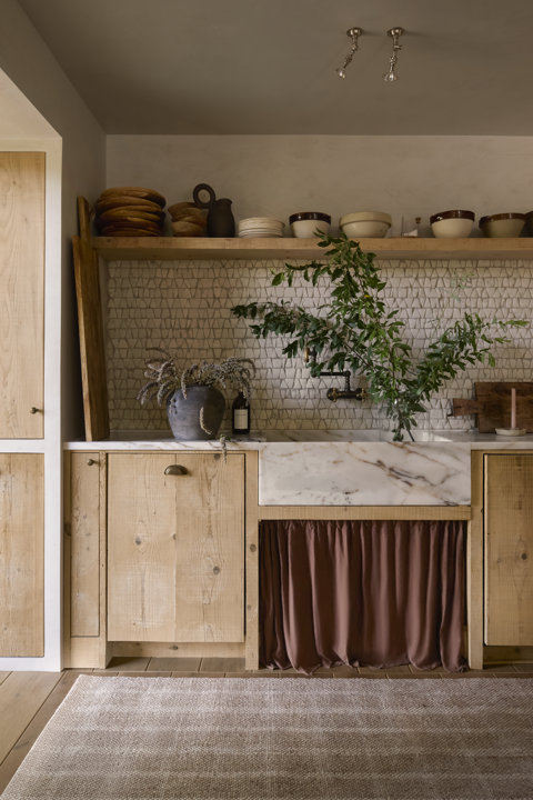 Cuisine dotée d'armoires en bois rustique, d'un évier de ferme en marbre avec une jupe en tissu brun et d'une étagère ouverte présentant de la poterie sur un dosseret de carreaux blancs texturés.