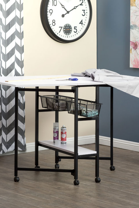 White sewing and craft table on wheels with black metal legs, storage baskets, and a lower shelf.