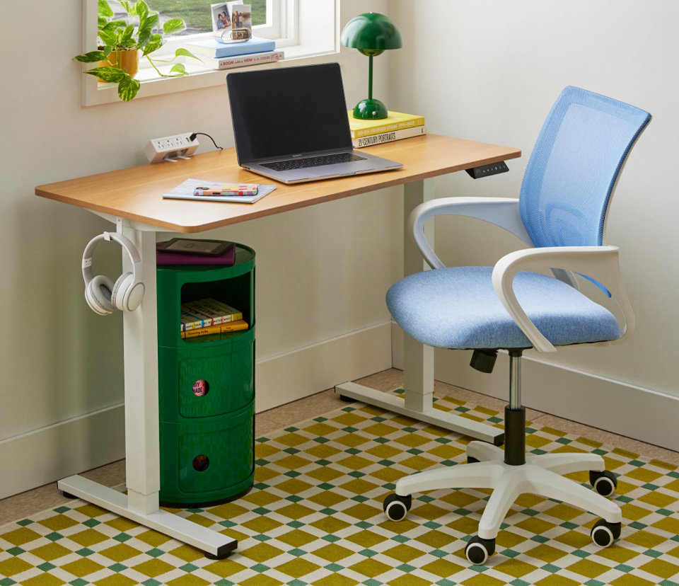 A wooden adjustable standing desk with a blue mesh chair, a laptop, and a green three-tier storage cart.