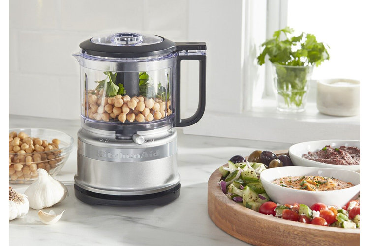 Stainless steel food processor filled with chickpeas and herbs next to a serving platter with bowls of hummus and vegetables.