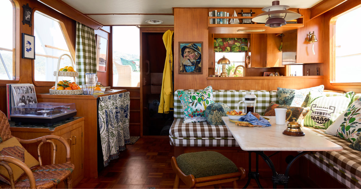 Cozy, rustic interior of a boat cabin featuring wood paneling, a marble-topped table with gingham bench seating, patterned throw pillows, a record player, and warm ambient lighting.