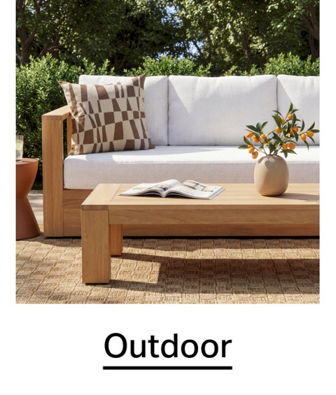 Outdoor patio furniture set featuring a teak sofa with white cushions, a matching coffee table with an open book, and a vase of citrus branches, all set on an outdoor rug.