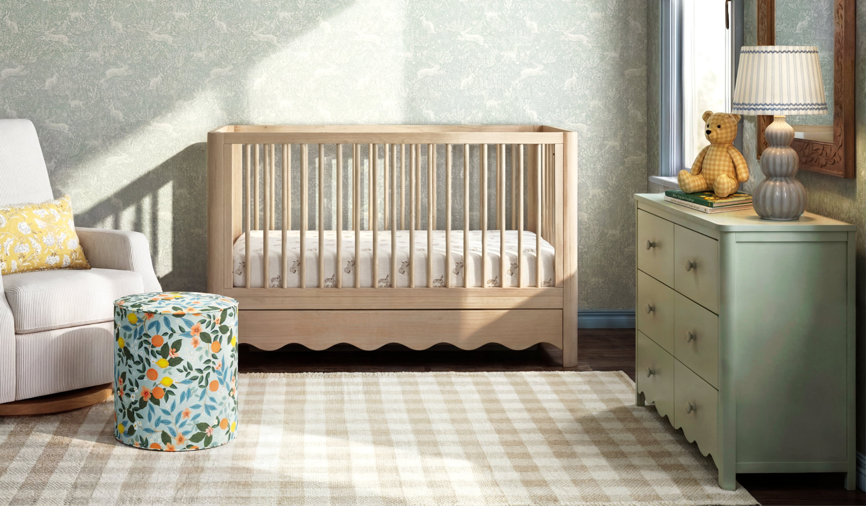 Light wood baby crib with a scalloped base in a nursery featuring sage green rabbit-patterned wallpaper, a gingham area rug, and a sage green dresser.