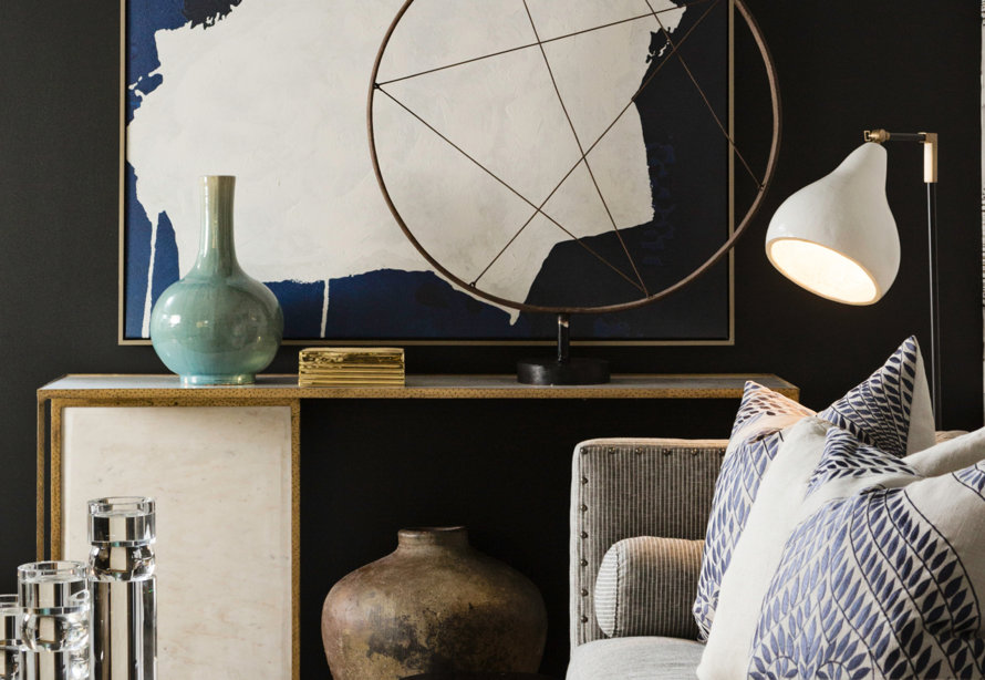 A console table featuring a marble base and gold trim sits against a black wall, topped with a teal vase, gold box, and an abstract metal circle sculpture, with a white task lamp and a patterned sofa in the foreground.