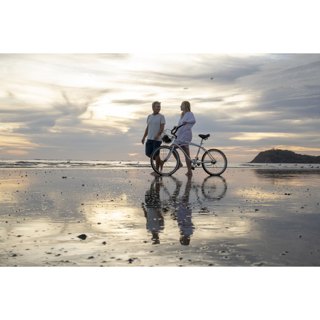 Älteres Paar fährt bei Sonnenaufgang mit dem Fahrrad am Strand entlang von AscentXmedia - Kein Rahmen Druck auf Leinwand