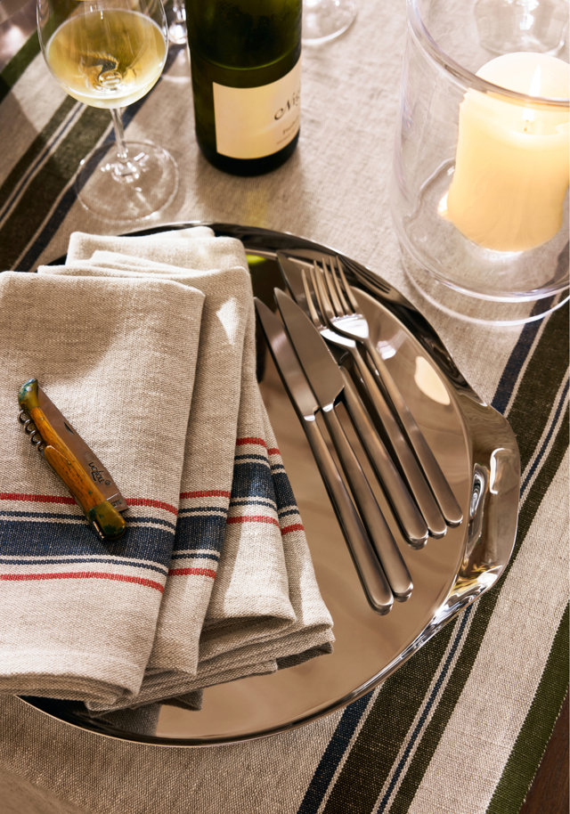 Table setting featuring a polished silver charger plate, stainless steel flatware, and a folded linen napkin with red and blue stripes, accompanied by a glass of white wine, a wine bottle, and a pillar candle in a glass hurricane.