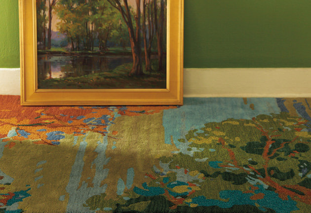 An artistic rug featuring a scenic impressionist-style landscape pattern with vibrant green, blue, and orange tones, placed next to a framed landscape painting against a green wall