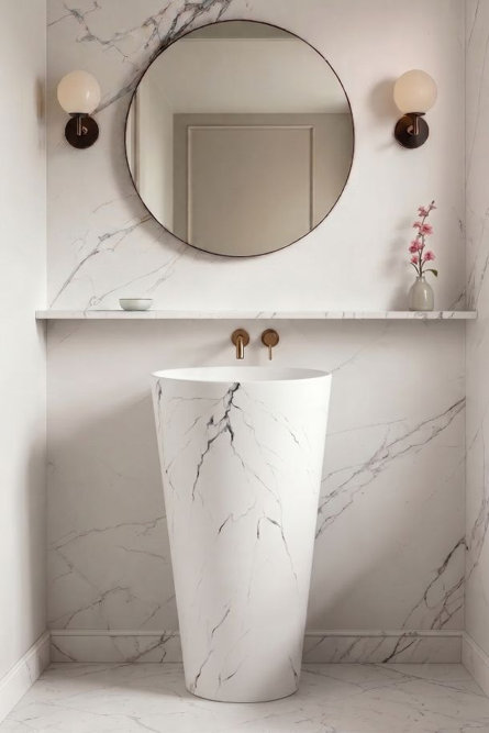 Modern pedestal sink with white and gray marble veining set against a matching marble tiled wall featuring a circular mirror and twin wall sconces.