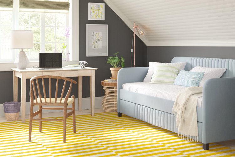 home office with a light blue daybed styled as a sofa with throw pillows beside light wood desk over a yellow geometric rug.