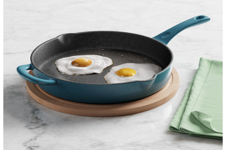 Blue frying pan with two fried eggs inside. Wood trivet on a marble countertop next to green fabric napkins