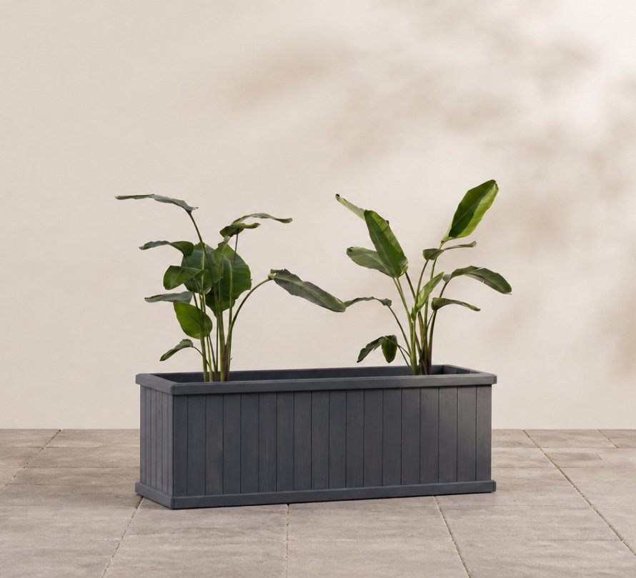 Two green leafy plants potted in a rectangular charcoal grey slatted wooden planter set on a stone tiled patio.