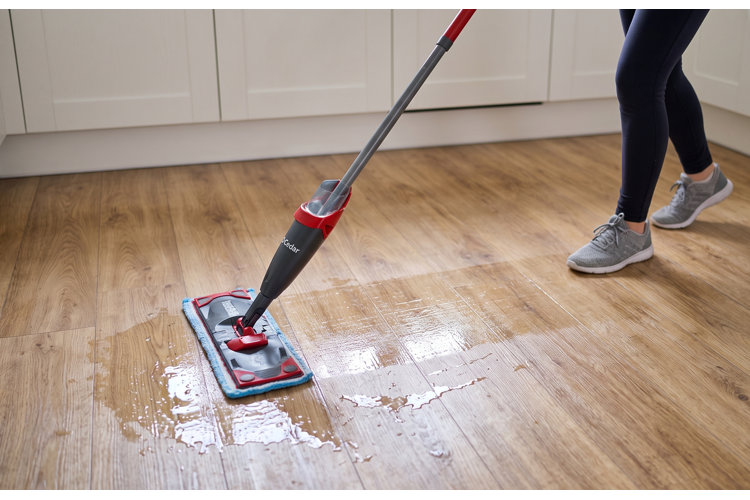 An action shot of a person mopping a light brown laminate floor with a spray mop. The person, wearing dark leggings and gray sneakers, is walking forward while using the red and gray mop, which leaves a visible trail of wet, clean sheen on the floor. In the background, white kitchen cabinets line the base of the wall.
