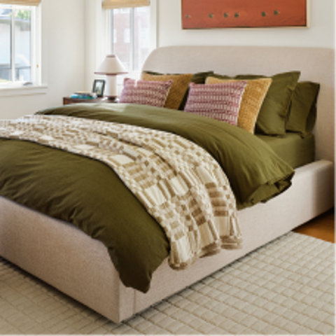 Beige upholstered bed frame with olive green bedding, textured throw blanket, and patterned accent pillows in a sunlit bedroom.