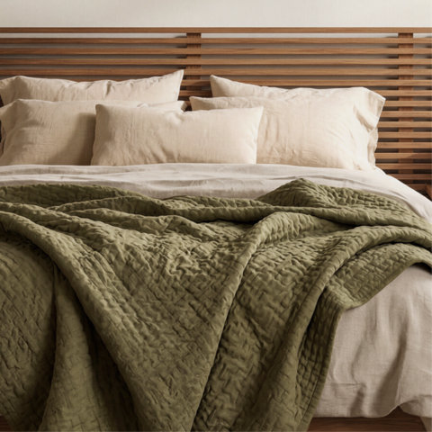 A cozy bed with a slatted wood headboard, layered with cream-colored linen pillows and a textured olive green quilt.