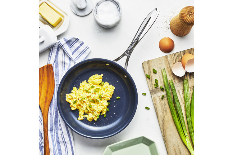 A navy blue frying pan with scrambled eggs and chopped scallions and surrounded by cracked egg shells and a wood spatula.