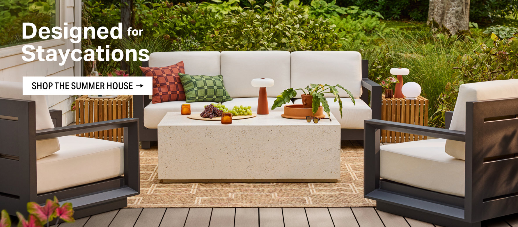 Designed for Staycations, shop the summer house with an outdoor seating area featuring a white sofa and two chairs arranged around a rectangular stone coffee table on a wooden deck.