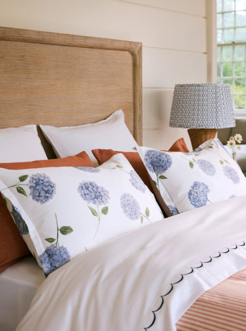 A cozy bedroom featuring hydrangea-print bedding and accent pillows, a wooden headboard, a white scalloped duvet, and a wicker lamp on a nightstand.