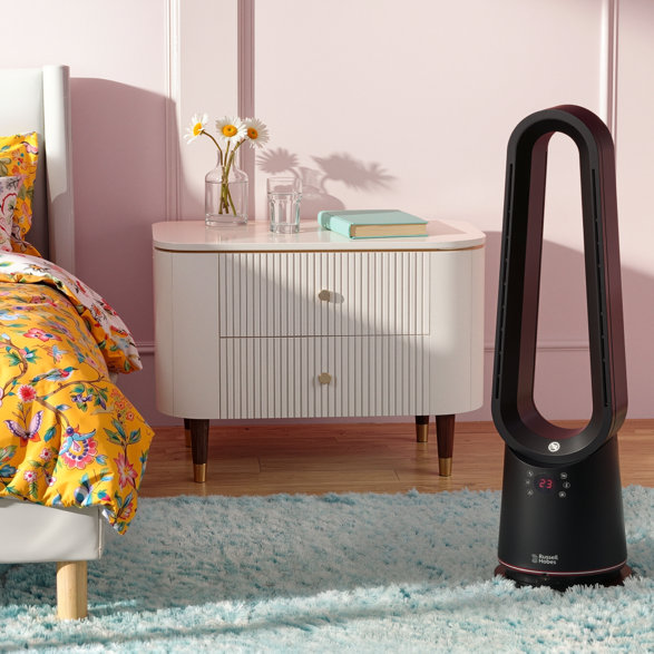 Black Russell Hobbs bladeless tower fan standing on a blue fluffy rug in a bedroom next to a white bedside table with a floral bedspread.