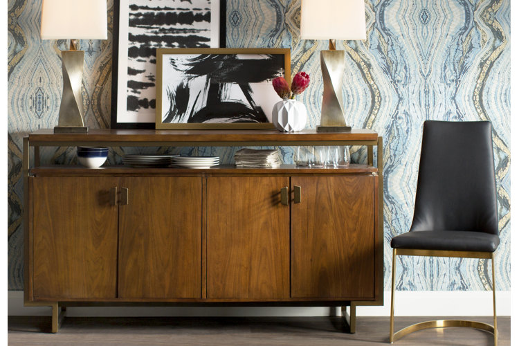 A stylish dining area featuring a mid-century modern wooden sideboard with a brass metal frame set against a dramatic blue, teal, and gold patterned wallpaper. The top surface of the credenza is decorated with two modern twisted brass lamps, framed black-and-white abstract art, and a geometric white vase holding red flowers. Underneath the main tabletop, an open shelf stores stacked plates and glassware, and a black leather chair with a brass metal base sits to the right.