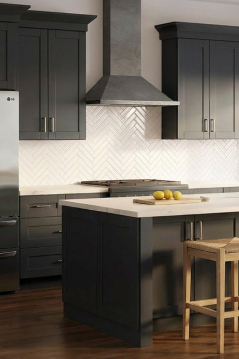 Modern kitchen with dark gray shaker cabinets, white herringbone backsplash, marble countertops, stainless steel appliances, and a wooden bar stool at the island.
