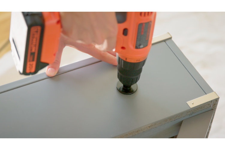 Person using an orange power drill with a circular drill bit to drill a hole into the back of a gray wood nightstand.
