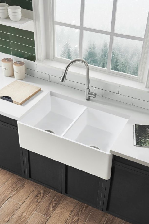 White double-bowl farmhouse kitchen sink with a stainless steel faucet installed in a modern kitchen with black cabinets and white countertops.