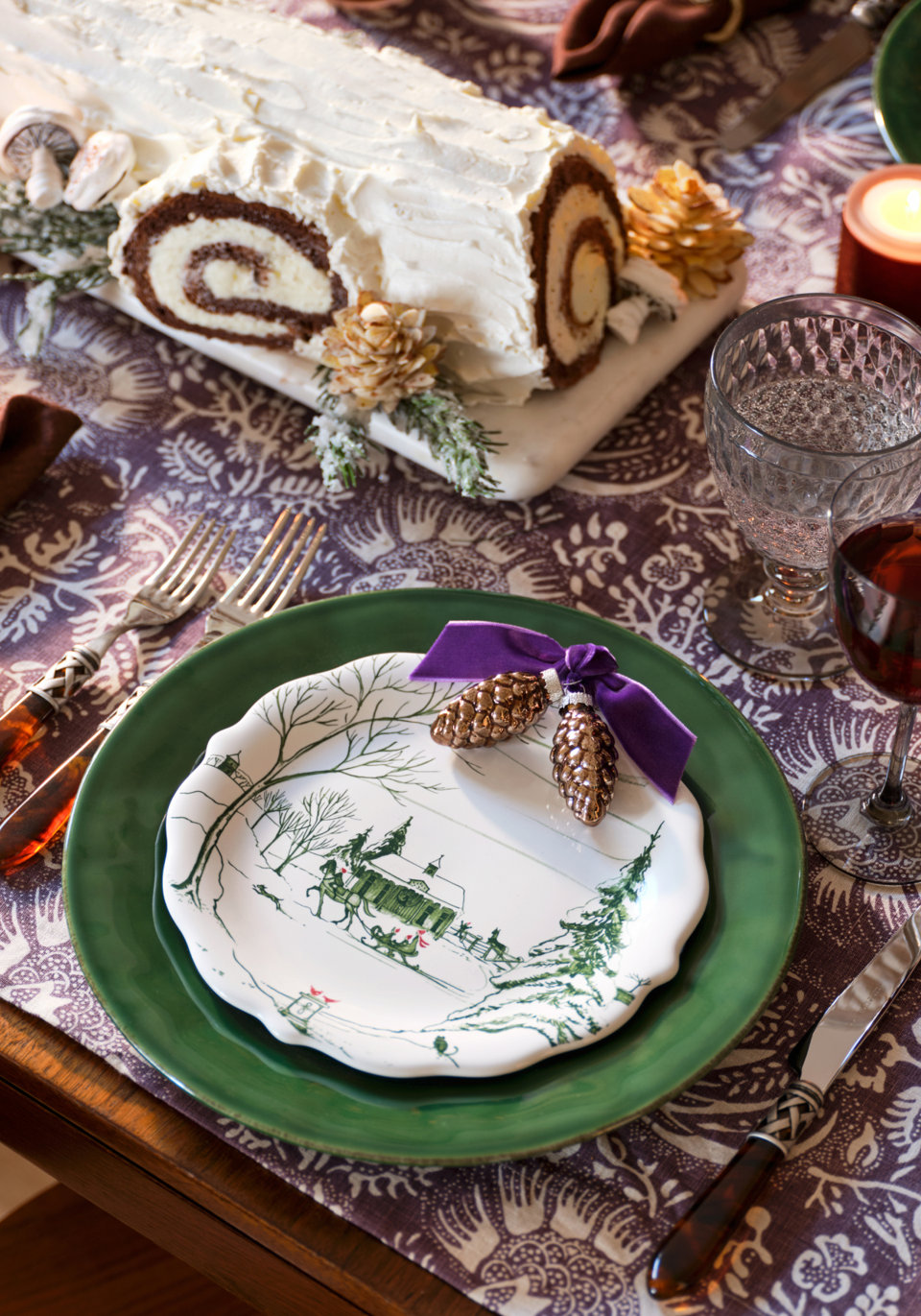 Festive holiday place setting with a green dinner plate, a salad plate featuring a winter scene, a Yule log cake, and an elegant patterned table runner.