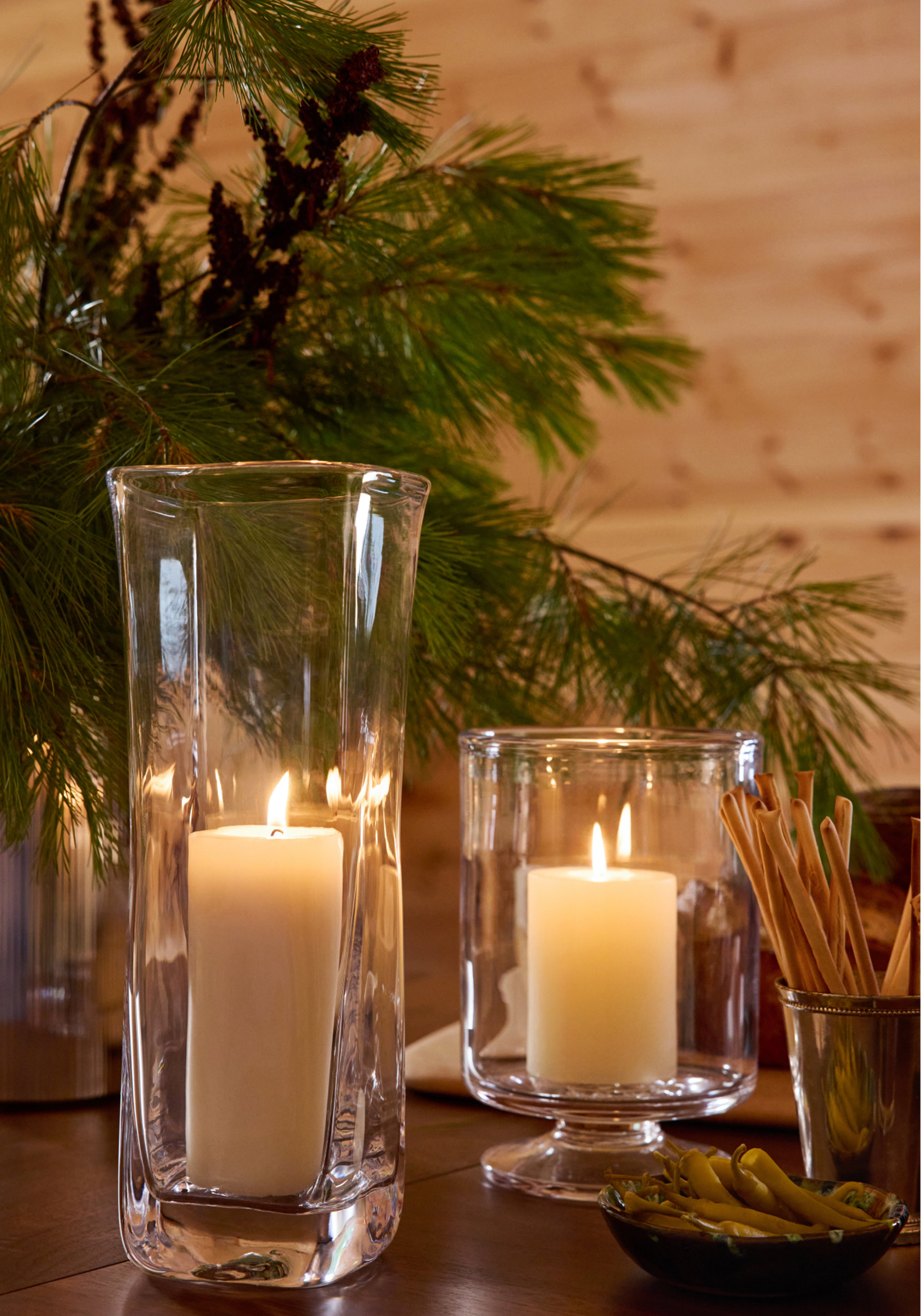 Two glass hurricane candle holders with lit white pillar candles arranged on a wooden table with pine branches, breadsticks, and a bowl of olives.