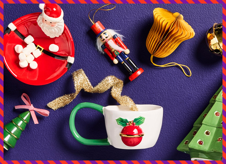 A festive flat lay on a dark purple background featuring a Santa-themed bowl filled with marshmallows, a nutcracker ornament, a gold paper bell, a ceramic mug with a holiday ornament design, a green tree-shaped ornament, and a gold ribbon.