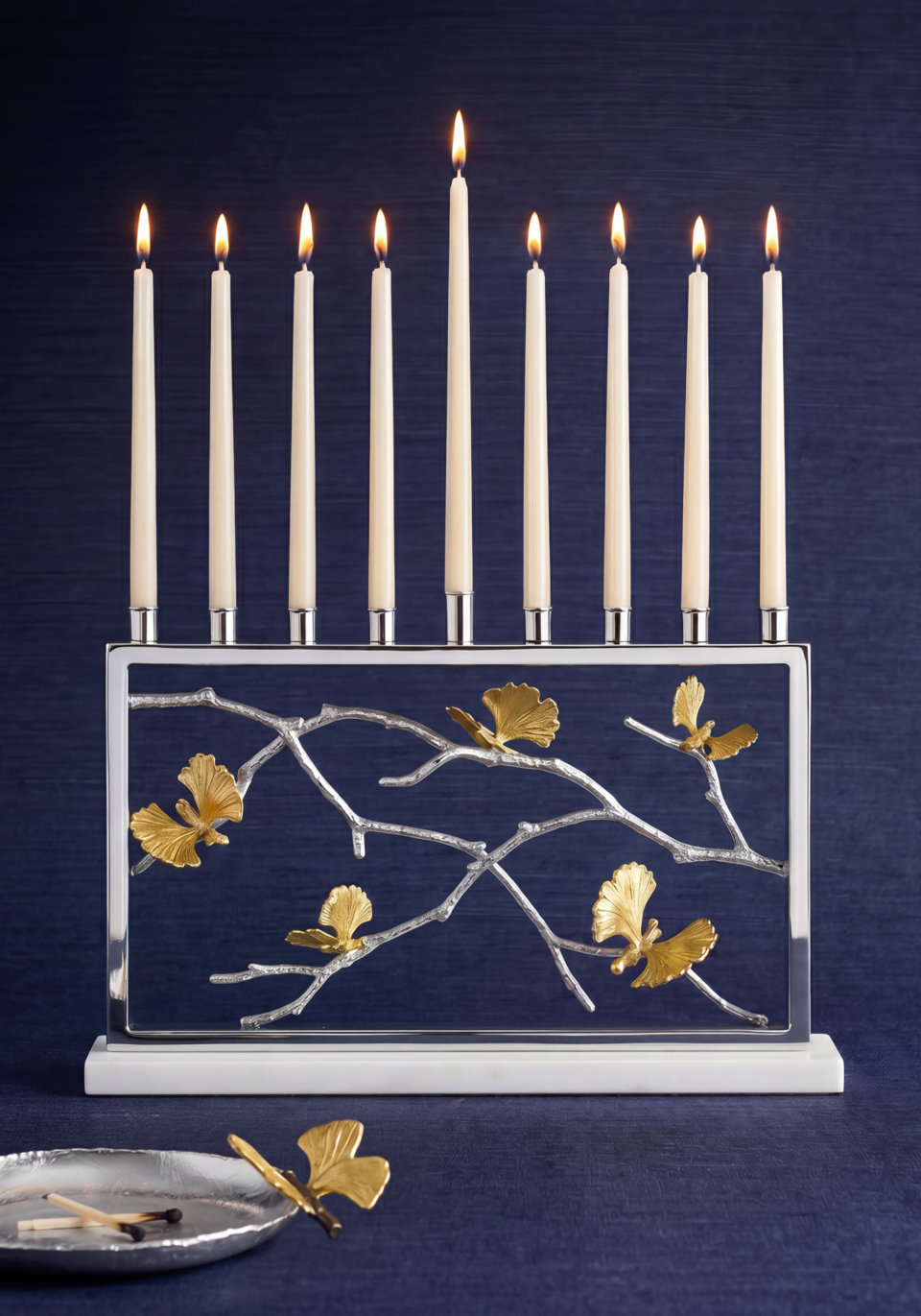 Silver menorah with gold ginkgo leaf accents and white candles lit on a blue background, with a small matching tray and matches in the foreground.