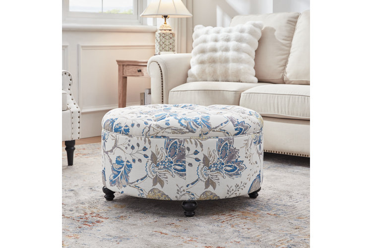 A large, round storage ottoman with a white fabric cover featuring blue and gray floral patterns, a button-tufted top, and dark wood turned bun feet, centered in a brightly lit living room