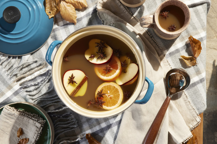 Tabletop with a plaid tablecloth under a blue dutch oven filled with sliced apples, oranges, cinnamon sticks, and cloves.