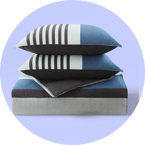 Une couette pliée bleue, grise et noire empilée avec deux oreillers assortis sur un fond violet.