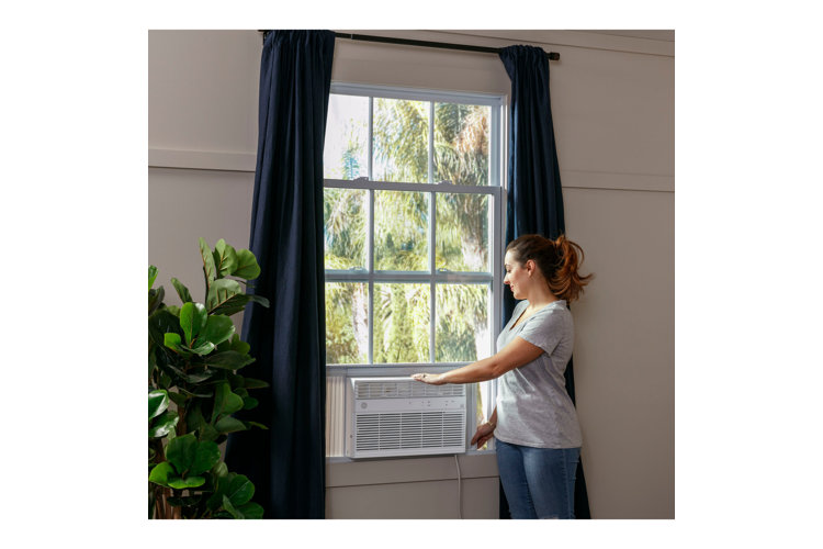A woman adjusting an AC unit in a window with long navy blue curtains and a large potted plant next to it