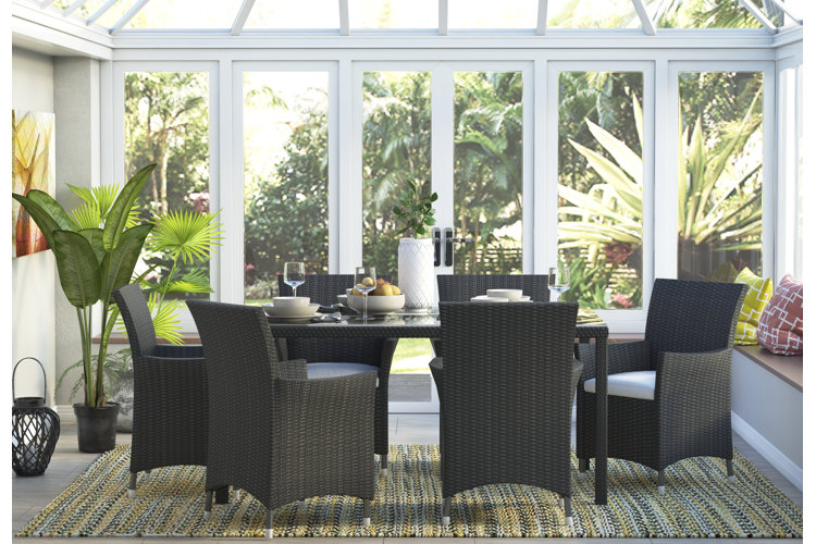 Wicker dining set with glass-top table and cushioned chairs in screened in porch with large windows, tropical plants, and outdoor greenery views.