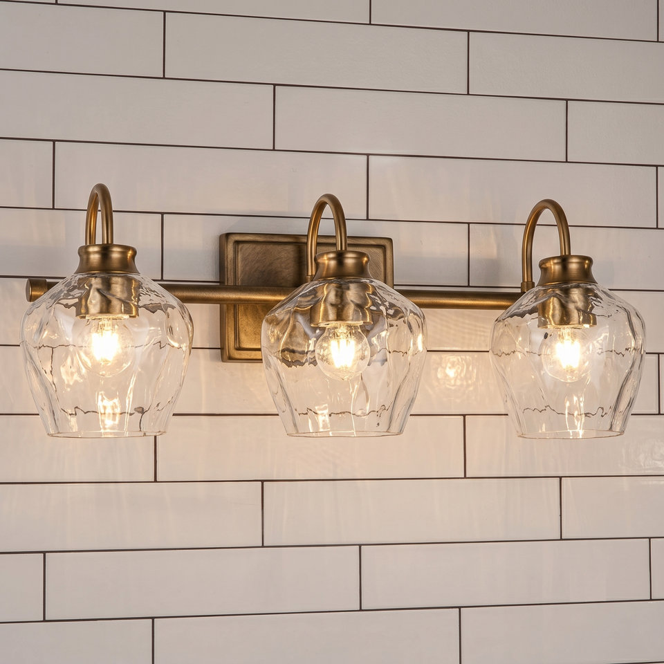 Three-light bathroom vanity fixture with gold-toned hardware, clear hammered glass shades, and a black mounting plate installed on a white subway tile wall above a mirror.