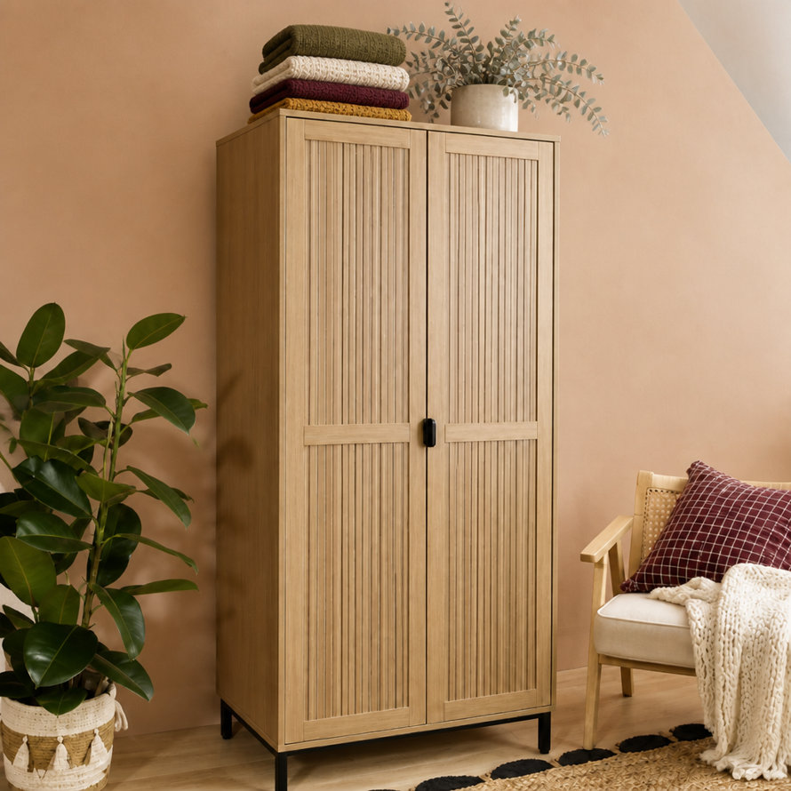 Tall light oak wardrobe with vertical slats and black metal legs, styled in a room with a potted rubber plant, a chair with a patterned cushion, and folded blankets on top.