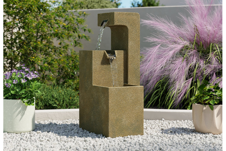 Patio with a square, three-tiered fountain surrounded by foliage and grass plants and placed on top of white gravel.
