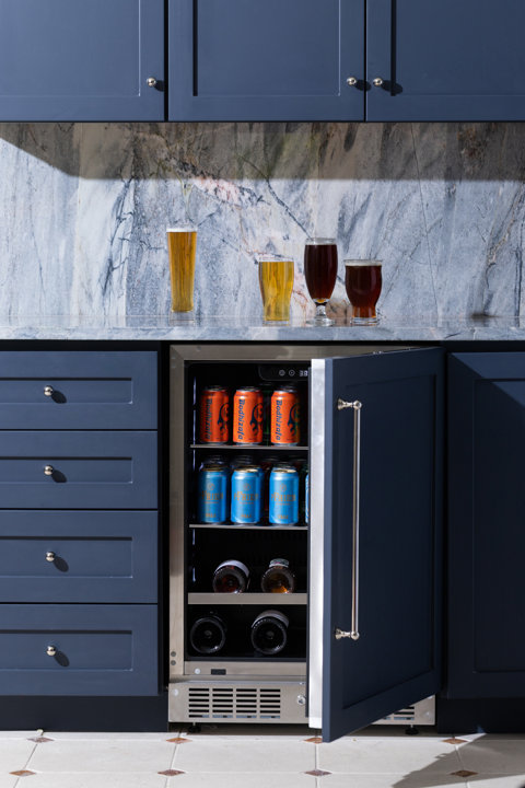Under-counter beverage refrigerator integrated into dark blue kitchen cabinetry with four glasses of beer on a marble countertop above.