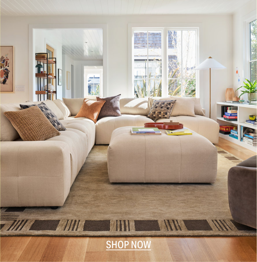 Beige modular sectional sofa with an oversized ottoman in a bright, sunlit living room with an area rug. SHOP NOW.