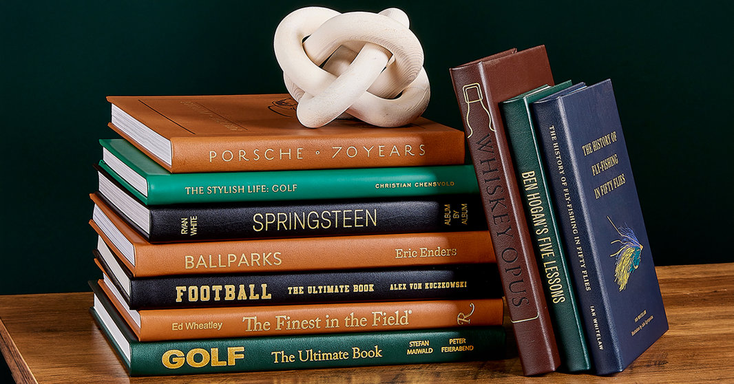 Stack of hardcover coffee table books on a wooden surface topped with a white decorative knot sculpture against a dark green background.