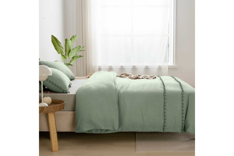Bed with a light green comforter and pillow set with embroidered pom-poms beside a window with white sheer curtains.