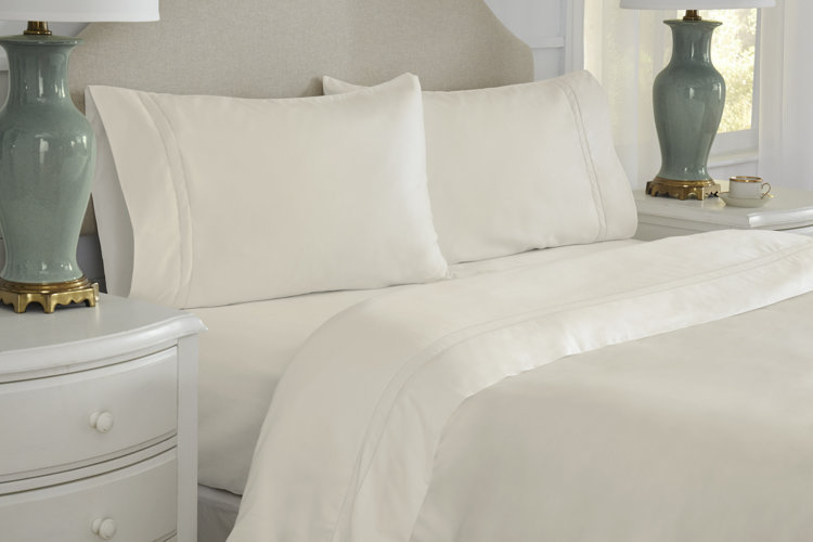 A cream sheet set on a bed with an upholstered headboard with green table lamps next to the bed