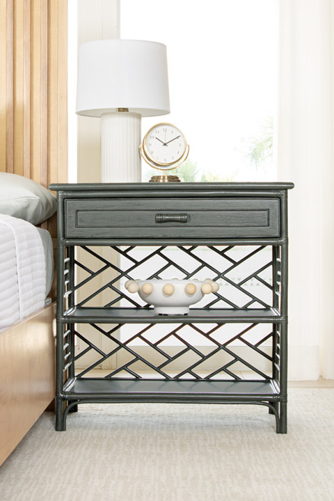 Dark gray rattan nightstand with a single drawer and two open shelves featuring a geometric lattice pattern, styled with a table lamp, clock, and decorative bowl.