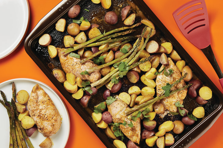 black sheet pan with chicken potatoes asparagus and herbs next to a red spatula and a dish with a serving of food